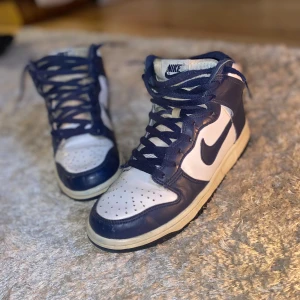 Nike Dunk High Navy Blue EUR 39 - Nike Dunk High sneakers i vit och marinblå färg med klassisk Swoosh-logga på sidan. Skorna har snörning, perforerad tå och hög siluett. Ovandel i skinn och textil, platt sula och rund tå. Perfekta för dig som gillar retrovibbar och streetstyle.