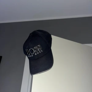 marinblå keps från Hugo Boss - Snygg mörkblå keps från Hugo Boss med tydlig broderad logga framtill. Klassisk böjd skärm och justerbar passform. Perfekt för dig som vill ha en stilren accessoar med premiumkänsla och diskret branding.
