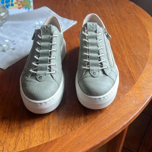 Gröna sneakers från Rieker med leoparddetalj - Snygga gröna sneakers från Rieker med vit plattsula, snörning och dragkedja på sidan. Baktill finns en cool leopardmönstrad detalj som ger extra edge. Skorna är i skinnimitation och har rund tå. Perfekta för dig som vill ha något unikt till vardagslooken.