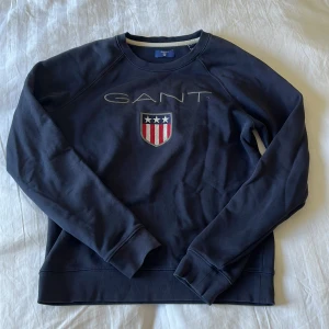 Mörkblå sweatshirt från GANT - Mörkblå sweatshirt från GANT med broderad logga och sköld med stjärnor och ränder på bröstet. Klassisk rund halsringning och långa ärmar. Tillverkad i mjuk bomull, perfekt för en chill och stilren look.