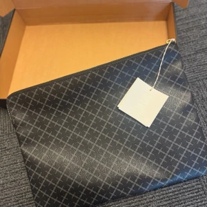 Datafoderal By Malene Birger - Stilren helt ny svart datafoderal från By Malene Birger med grått rutigt mönster och guldfärgad dragkedja. Väskan är tillverkad i syntetmaterial och har ett ljust innerfoder samt en praktisk innerficka. Perfekt för att bära laptop eller viktiga papper.