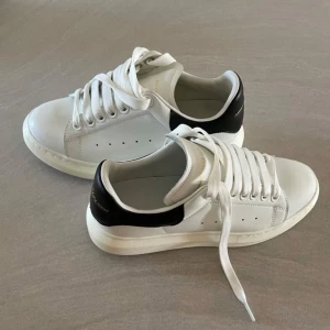 Alexander McQueen vita sneakers - Stilrena vita sneakers från Alexander McQueen med svart häl och tjock sula. Skorna har klassisk snörning, rund tå och är tillverkade i skinn. Perfekta för dig som gillar en clean och modern look med lyxig känsla. Endast använda en gång!