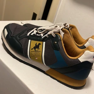 U.S. Grand sneakers i grå och gul - Snygga sneakers från U.S. Grand Polo Equipment & Apparel med grå mesh, mörkblå och vita detaljer samt gul insida. Skorna har vit snörning, svart sula och coolt logomärke på sidan. Perfekta för dig som gillar sportig och stilren look.