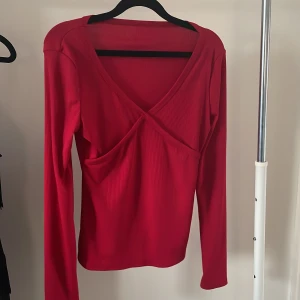 Röd omlottopp med ribbad struktur - Snygg röd omlottopp med ribbad struktur och lång ärm. Toppen har en djup v-ringning och omlottdetalj framtill. Materialet är mjukt och stretchigt. Har blivit använd endast en gång, prislappen är inte kvar. 