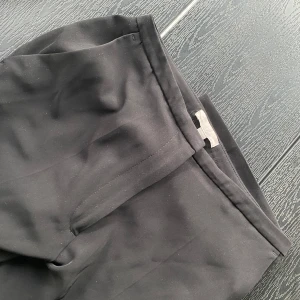 Svarta kostymbyxor med raka ben - Snygga svarta kostymbyxor med raka ben och diskreta fickor bak. Klassisk design med slät front och stilren look, perfekta och trendiga nu till hösten. Materialet är stilfullt och har en lätt glansig finish. Sparsamt använda.