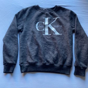 Mörkgrå sweatshirt från Calvin Klein - Säljer en mörkgrå sweatshirt från Calvin Klein Jeans med stor vit CK-logga framtill. Tröjan har rund hals, långa ärmar och ribbade muddar vid ärmslut och nederkant. Perfekt för en chill och stilren look. L men passar M