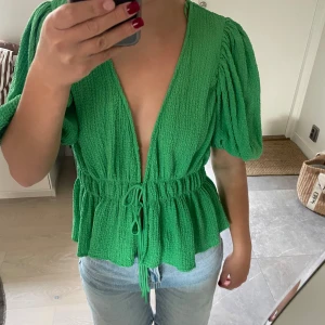 Grön blus med puffärm från Zara - Superfin grön blus från Zara med djup v-ringning, puffiga korta ärmar och knyt i midjan. Blusen har en strukturerad, lite krinklad yta och ger en luftig och trendig look. Perfekt till jeans eller kjol för en färgstark outfit.
