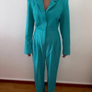 Turkos kostym jumpsuit med axelvaddar - Snygg turkos jumpsuit med långa ärmar och markerade axelvaddar. Djup v-ringning och klassisk krage ger en cool och stilren look. Byxdelen är rak och materialet känns lätt och smidigt. Perfekt för dig som vill sticka ut med färg och form.