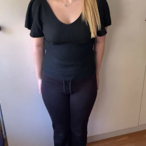 Svart ribbad topp med puffärm - Svart ribbad topp med v-ringning både fram och bak samt korta puffärmar. Toppen har en snygg knytning i nacken och sitter figurnära. Perfekt för dig som gillar en enkel men ändå trendig look.