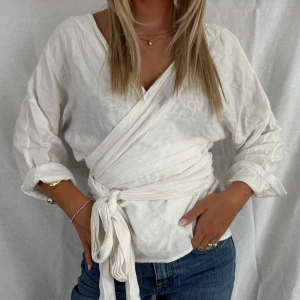Vit omlottblus i linne - Snygg vit omlottblus med knyt i midjan och v-ringning. Blusen har långa ärmar som kan rullas upp och är gjord i ett luftigt material som påminner om linne. Perfekt till jeans för en chill och stilren vibe.