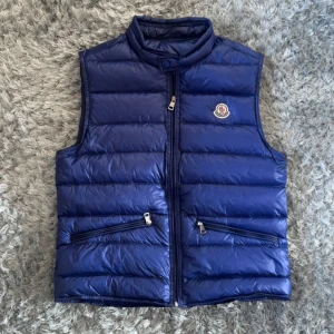 Blå dunväst från Moncler - Säljer en blå dunväst från Moncler med hög krage och två snedställda fickor med dragkedja. Västen har quiltad design och klassisk Moncler-logga på bröstet. Perfekt för lager-på-lager och riktigt snygg till streetwear.