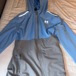 Blå och grå vindjacka från Under Armour - Snygg tvåfärgad vindjacka från Under Armour i blått och grått. Jackan har huva, hel dragkedja och reflekterande detaljer på axlarna. Perfekt för blåsiga dagar och har praktiska fickor med dragkedja. Sportig look och lätt material.