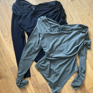 Asymmetrisk långärmad body - Två snygga långärmade body med asymmetrisk one shoulder-skärning och draperad effekt. En i svart och en i olivgrön, båda i mjukt stretchigt material. Perfekt för dig som vill sticka ut med coola detaljer och unik passform.