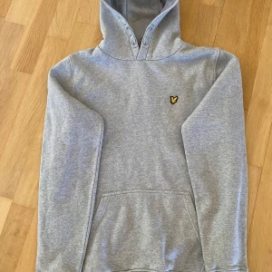 Grå hoodie från Lyle & Scott Junior - Snygg grå hoodie från Lyle & Scott Junior med klassisk gul logga på bröstet. Tröjan har huva, känguruficka och långa ärmar. Perfekt för en chill och avslappnad stil.