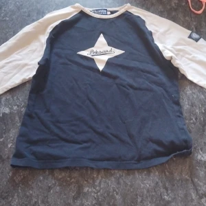 Leksand långärmad tröja blå/vit - Snygg långärmad tröja från Leksand Official Wear i marinblått med vita ärmar. Stor logga med texten 'Leksand' på bröstet. Perfekt för dig som vill visa ditt stöd för laget och gillar sportig stil.