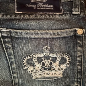 Victoria Beckham Rock & Republic jeans - Snygga blå jeans från Victoria Beckham för Rock & Republic med raka ben och broderade kronor på bakfickorna. Jeansen har en klassisk tvätt och är tillverkade i jeansmaterial. Perfekta för dig som vill sticka ut med coola detaljer.