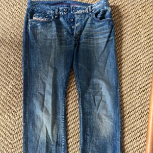 Blå jeans från Diesel, storlek 34 - Säljer ett par klassiska blå jeans från Diesel i storlek 34. Jeansen har snyggt slitna detaljer, raka ben och knäppning med knappar. De är tillverkade i bomull och har den ikoniska Diesel Industry-loggan på myntfickan. Perfekta för en avslappnad och cool stil.