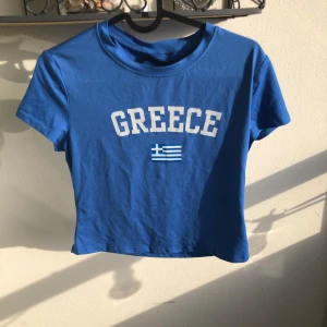 Blå croppad t-shirt  - En blå croppad t-shirt med vit text 'GREECE' och en liten grekisk flagga på bröstet. Jätte skönt tyg i bomull.