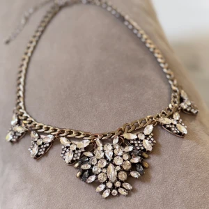 Statement halsband med glittriga stenar - Snyggt statement halsband med tjock guldfärgad kedja och stora glittriga stenar i olika former. Stenarna är klara och sitter i blommiga mönster längs kedjan. Perfekt för att lyfta vilken outfit som helst med extra bling.