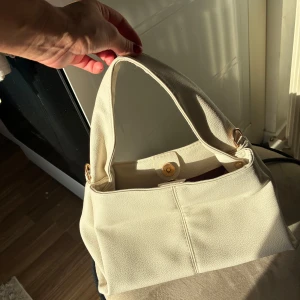 Krämvit axelväska i skinnimitation - Snygg axelväska i krämvitt med enkel, stilren design och bred axelrem. Väskan har magnetlås i guldton och vinrött innerfoder. Perfekt storlek för vardagens essentials och lätt att matcha till olika outfits.