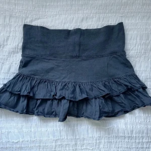 Mörkgrå volangkjol med fjäril - Supersöt mörkgrå kortkjol med dubbla volanger och bred midja. Liten rosa fjäril broderad framtill som detalj. Kjolen är i mjuk bomull och passar perfekt till sneakers eller boots för en chill look.