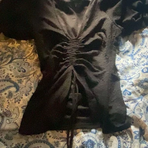 Svart snörad topp från BikBok XS - Supersnygg svart topp från BikBok i storlek XS. Toppen har en cool snörningsdetalj framtill som ger en rynkad effekt och är gjord i ett strukturerat, lite glansigt tyg. Kortärmad och figurnära passform, perfekt att styla med jeans eller kjol.