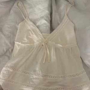 Krämvit linnetopp från Stradivarius - Supersöt krämvit linnetopp från Stradivarius med smala axelband, v-ringning och spetsdetaljer. Toppen har knyt framtill och smockad rygg för extra komfort. Perfekt för varma dagar och ger en romantisk vibe till din outfit.