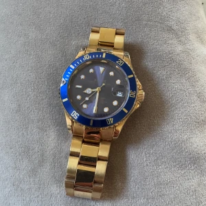 Guldig klocka med blå urtavla - Snygg guldig klocka med blå urtavla och matchande blå ring runt glaset. Klockan har datumvisning och tydliga indexmarkeringar. Länken är i metall och ger en lyxig känsla. Perfekt accessoar för dig som vill sticka ut.