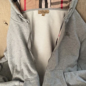 Grå zip  från Burberry - Snygg grå zipfrån Burberry med klassiskt rutigt foder i huvan. Jackan har dragkedja framtill, två fickor och är fodrad med mjuk fleece. Perfekt för dig som vill ha en stilren och bekväm jacka med exklusiv känsla.dragkedjan är trasig så den går inte att stänga.