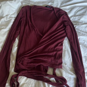 Vinröd omlottopp från 157 - Säljer en vinröd omlottopp från 157 i mjukt material. Toppen har långa ärmar, v-ringning och knytband i midjan för en snygg passform. Perfekt för dig som gillar stilrena och trendiga plagg med lite extra detalj. Har endast använt den 1 gång så är ej sliten alls.