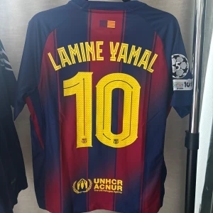 FC Barcelona Lamine Yamal #10 fotbollströja - Säljer en FC Barcelona matchtröja med Lamine Yamal och nummer 10 på ryggen. Tröjan är blå och röd med gula detaljer, klubbmärke, Nike-logga och sponsorer som Spotify och UNHCR. Kortärmad modell i lätt och ventilerande material, med Champions League-märke på ärmen.