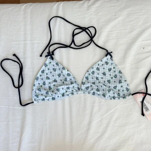 Blommig blå bikiniöverdel från Nelly - Supersöt trekantsbikini från Nelly med ljusblå och vit rutig bas och små blå blommor. Svarta knytband i hals och rygg samt en liten rosett upptill. Perfekt för stranden och sommarens alla dopp!