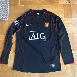 Manchester United Ronaldo 7 långärmad tröja - Säljer en svart långärmad Manchester United fotbollströja från Nike med Ronaldo 7-tryck på ryggen. Tröjan har röda detaljer, Champions League-märke på ärmen och AIG som sponsor på bröstet. Materialet är lätt och andas, perfekt för match eller träning.