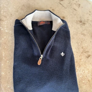 Mörkblå merinoullströja från Morris - Snygg mörkblå långärmad tröja från Morris i 100% merinoull. Tröjan har half zip med läderdetalj på dragkedjan och en broderad logga på bröstet. Perfekt för dig som gillar stilrena och klassiska plagg med en modern touch.