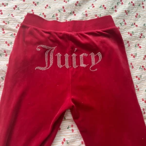 Röda velourbyxor Juicy Couture S - Röda velourbyxor från Juicy Couture med strassdetaljer och Juicy-text baktill. Byxorna har snörning i midjan, raka ben och JC-logga framtill. Perfekta för en chill och trendig look.