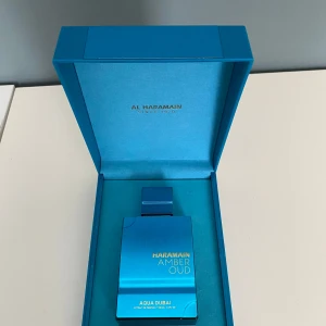 Al Haramain Amber Oud Aqua Dubai - Det är ungefär 95 ml kvar. Exklusiv parfym i en stilren, metallisk blå flaska med matchande lyxig box. Flaskan har en fyrkantig form och guldiga detaljer. Doften är Amber Oud Aqua Dubai, vilket ger en känsla av fräschör och elegans. Perfekt för dig som gillar unika och sofistikerade dofter.  
