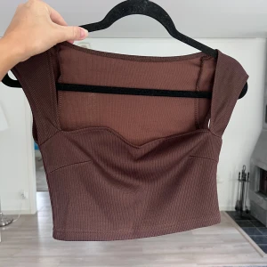 Brun croppad topp - Säljer en brun croppad topp i ribbat material. Toppen har breda axelband och en snygg sweetheart-urringning framtill. Tillverkad i stretchigt tyg som sitter tajt och bekvämt. Perfekt att styla med höga jeans eller kjol 🤎Önskar du veta exakta mått? Tveka inte att höra av dig!