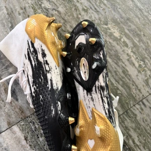 Adidas fotbollsskor i vitt, svart och guld - Snygga Adidas fotbollsskor med högt skaft i vitt, svart och guld. Skorna har coolt mönster, guldiga dobbar och klassiska tre ränder på sidan. Ovandelen är i syntetmaterial och snörning framtill. Perfekta för dig som vill sticka ut på planen.