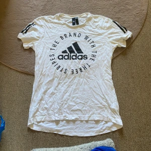 Adidas T-shirt  - Vit t-shirt från Adidas med svart logga och texten 'The brand with the three stripes' i cirkel på bröstet. Klassiska svarta ränder på axlarna. Skön passform och tillverkad i mjuk bomull. Perfekt för en sportig look!🙌🏻