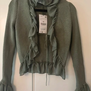 Olivgrön volangkofta från Zara - Supersöt olivgrön kofta från Zara med ribbad struktur och volangdetaljer längs kanterna och ärmsluten. Croppad modell med knyt framtill och långa ärmar. Perfekt för dig som gillar romantiska vibes och trendiga detaljer.