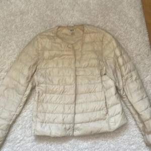 Beige dunjacka från Uniqlo XS - Lätt och smidig beige dunjacka från Uniqlo i storlek XS. Jackan har rund halsringning, horisontella sömmar och stängs med tryckknappar. Perfekt för lager-på-lager och enkel att packa ner i väskan.