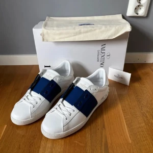 Valentino Garavani Open sneakers vit/blå - Snygga Valentino Garavani Open sneakers i vitt skinn med bred marinblå rand över vristen. Klassisk låg modell med snörning och ikoniska nitar på hälen. Perfekta för dig som vill ha en stilren men ändå unik look.
