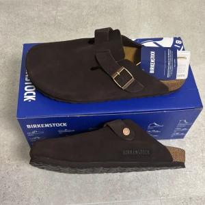 Birkenstock Boston bs bruna - Mörkbruna Birkenstock Boston sandaler i mjuk mocka med justerbart spänne i metall och klassisk korksula. Perfekt för dig som gillar en clean och chill look. Anatomisk fotbädd för extra komfort och snygg branding på sidan.