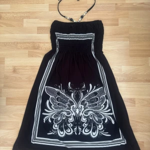 Svart och vit halterneckklänning - Svart långklänning med halterneck och snygga pärldetaljer i halsen. Klänningen har ett vitt, stort mönster framtill som påminner om fjärilsvingar och smock upptill för en bekväm passform. Perfekt för sommardagar när du vill sticka ut lite extra. Använd en gång.