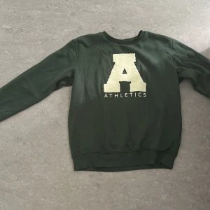 Grön sweatshirt med Athletics-tryck - Mörkgrön sweatshirt med stort vitt A och texten ATHLETICS framtill. Tröjan har rund hals, långa ärmar och ribbade muddar vid ärmslut och nederkant. Perfekt för en sportig och avslappnad stil.
