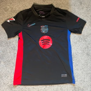 Barcelona fotbollströja - Lamine Yamal  - Snygg svart FC Barcelona fotbollströja från Nike med Lamine Yamal och nummer 19 på ryggen. Tröjan har röda och blå sidopaneler, klubbmärke och Spotify-logga framtill. Tillverkad i lätt och ventilerande material, perfekt för match eller träning. Storlek S.