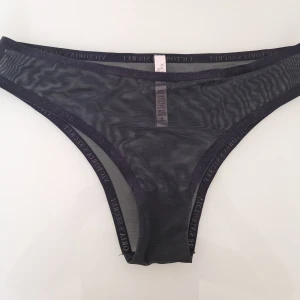 Svart Victoria's Secret trosa - Elegant svart trosa med spetsdetaljer. Den har en diskret Victoria Secret logga runt hela trosan och är tillverkad i ett mjukt och bekvämt material.