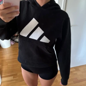 Svart Adidas hoodie med stor logga - Svart hoodie från Adidas i storlek S med stor vit logga framtill. Klassisk känguruficka, huva och långa ärmar. Perfekt för chill eller träning. Mjuk bomullsblandning som är skön att ha på sig.