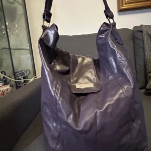 Lila axelväska från Daniel Hechter - En lila axelväska med stort huvudfack, dragkedja och innerfickor. Finns slitage, se bilder. Storlek: ca 33x30x12