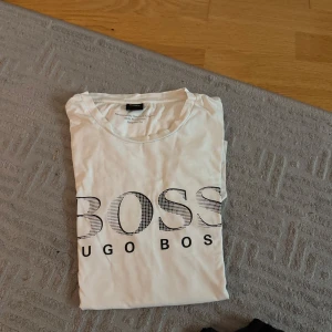 Vit t-shirt från Hugo Boss - Vit t-shirt från Hugo Boss med stor svart logga framtill. Klassisk rund halsringning och korta ärmar. Tillverkad i mjuk bomull, perfekt för en clean och stilren look.
